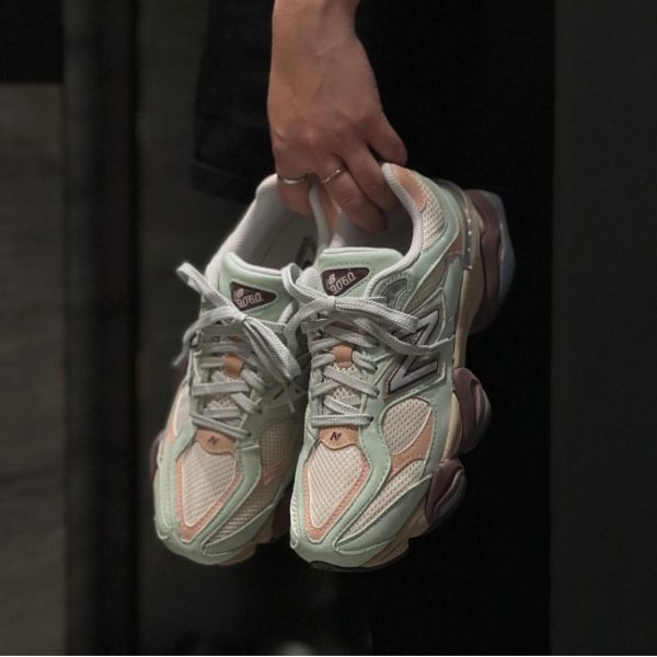 Person holding pastel mint and pink New Balance 9060 sneakers by the laces in front of a dark background, showing the side and toe details.