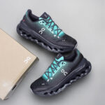 Pair of teal and black On running Cloud shoes with hollow pod sole, laid side by side beside a beige shoebox.