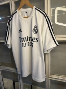 White Real Madrid soccer jersey on a wooden hanger, featuring the Emirates Fly Better sponsor and club crest.