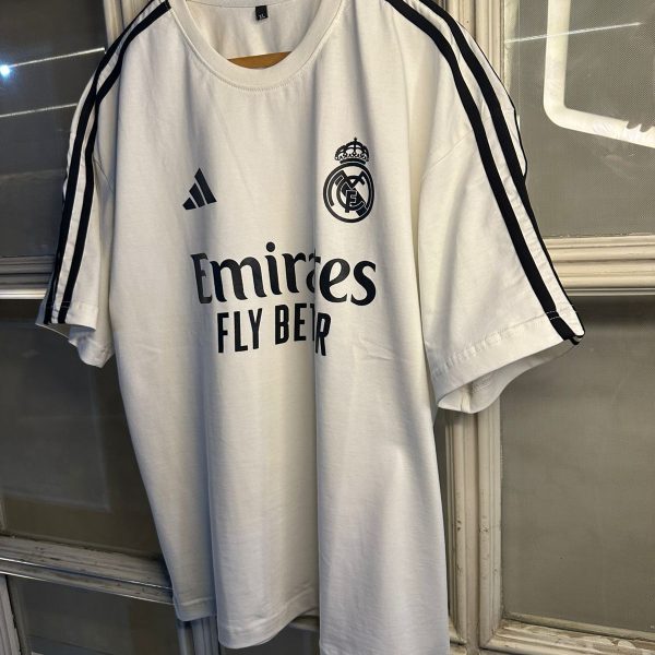 White Real Madrid soccer jersey on a wooden hanger, featuring the Emirates Fly Better sponsor and club crest.