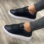 5c8b9efc-46bf-462a-aaeb-3d96cb5b63e9_3175e60c-7e99-45e1-8175-45521ad509de | UAE SPORT STORE Black leather sneakers with white soles worn with gray jeans, a delicate gold ankle bracelet visible on one ankle on a wood floor