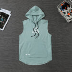 Mint-green sleeveless hoodie laid flat with a hood, black drawstrings, and a kangaroo pocket on a dark wooden surface.