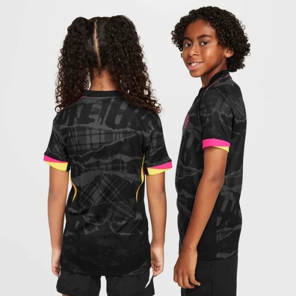 Two children showing the back and side of black graphic athletic jerseys with pink cuffs and yellow trim against a gray background, kid-focused fashion photo