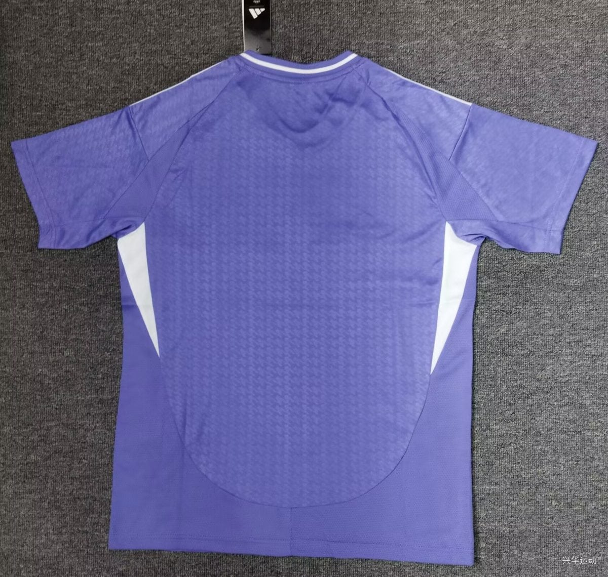 Purple athletic jersey hanging on a display hook with white side panels visible against a dark carpeted background, showing the back side and collar details.