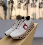 White running shoes with a red circular logo on the side, displayed on a beige box outdoors.