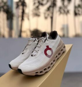 White athletic sneakers with a hollow outsole sits on a beige display box outdoors, featuring a red circular logo on the side.