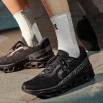 Black On running sneakers with a sculpted outsole, paired with white socks, standing on a paved surface.