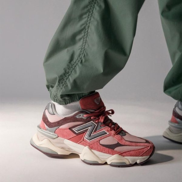 Close-up of a pink-red athletic sneaker with a chunky white sole, worn with sage green pants against a gray backdrop.