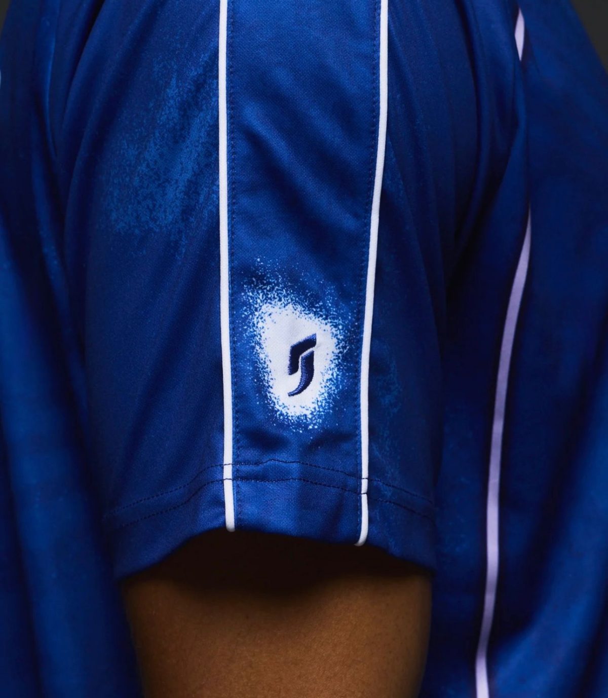 Close-up of a blue sports jersey sleeve with white piping and a white spray-painted circular emblem containing a dark abstract logo.