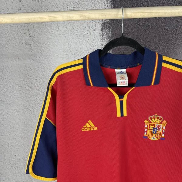 Red Spain national football jersey on a hanger, blue collar with yellow trim, crest on the chest and Adidas logo visible.