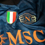 5l | UAE SPORT STORE Close-up of blue fabric with an Italian flag shield patch and a circular emblem featuring an orange N and skeletal hands.
