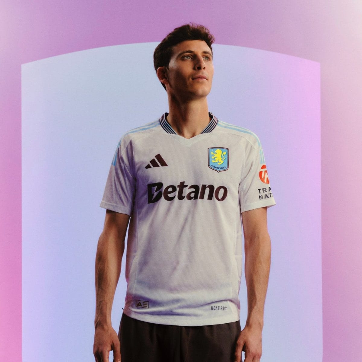 Young man in a white Aston Villa football jersey with Betano sponsor stands against a pink-purple gradient background, looking upward.