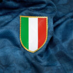 5m | UAE SPORT STORE Shield patch with the Italian flag (green, white, red) on textured blue fabric of a sports jersey, gold border visible.