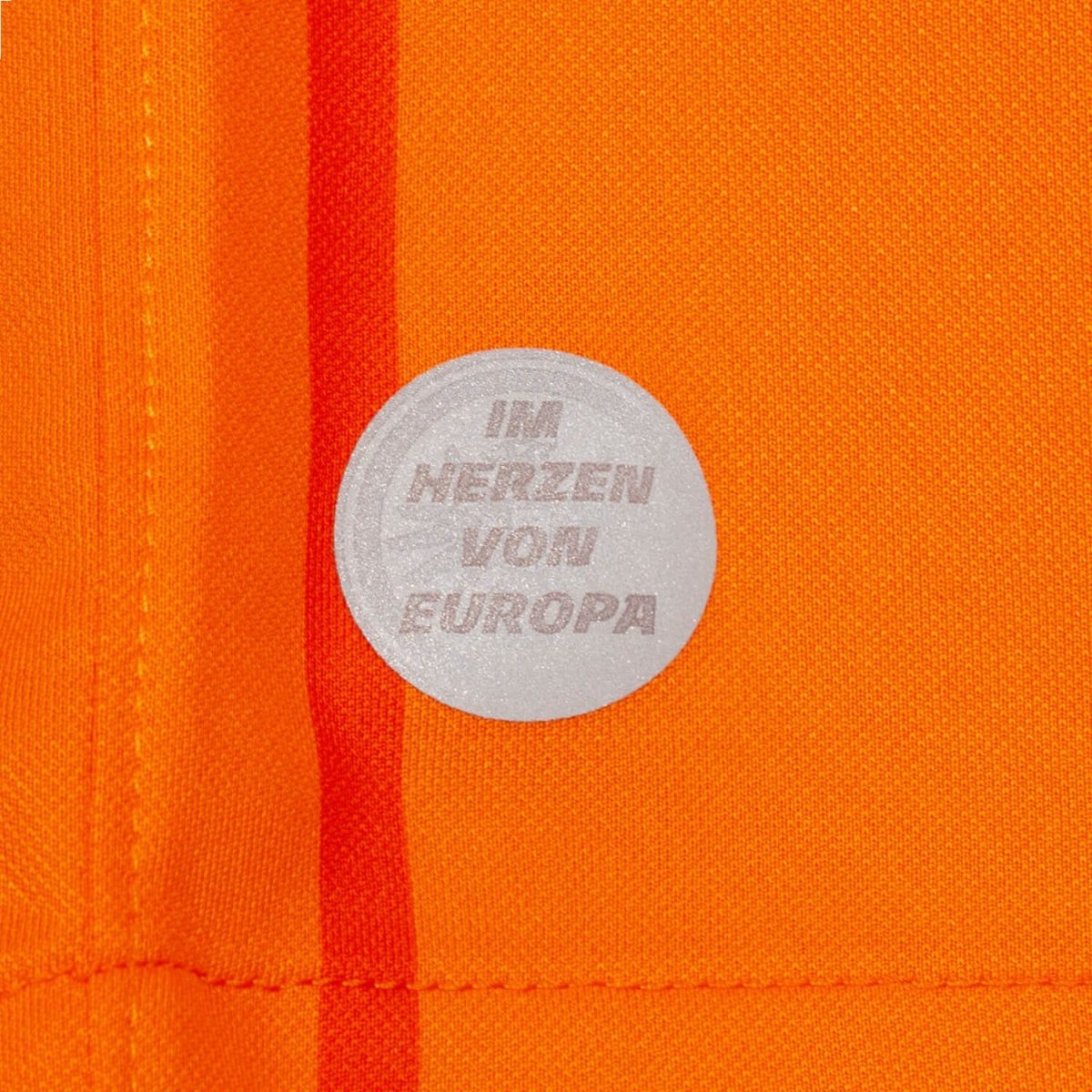Circular gray pin badge on bright orange fabric, text reads 'I AM HERZEN VON EUROPA'.