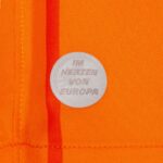 5sEmpORQALA4uBU | UAE SPORT STORE Circular gray pin badge on bright orange fabric, text reads 'I AM HERZEN VON EUROPA'.