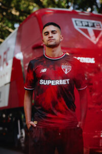 Man in a red and black São Paulo FC jersey with SUPERBET sponsor, standing with hands in pockets in front of a red team's van.
