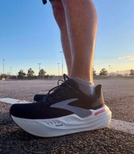 Close-up of a black Brooks running shoe with a white sole on textured pavement at sunset, worn by a person standing.