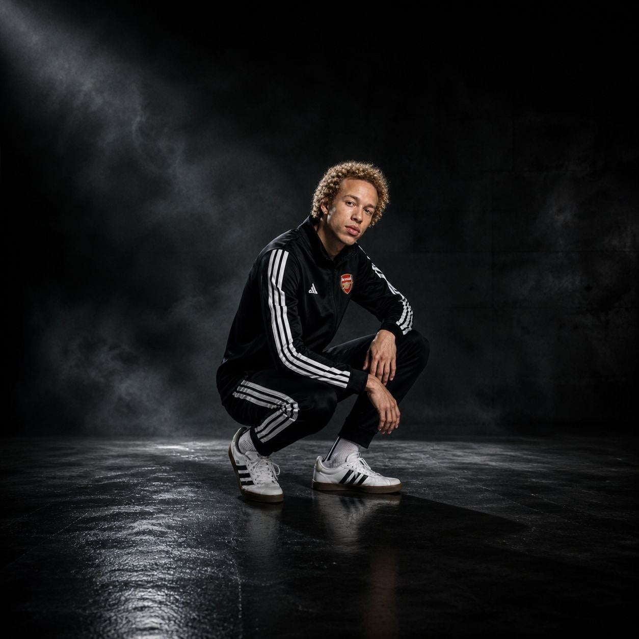 Athlete in a black Adidas tracksuit crouches on a dark studio floor with smoky lighting, wearing white sneakers.