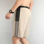 Beige athletic shorts with black side stripes and a black mesh panel on the side, worn by a person standing sideways with a wristwatch nearby.