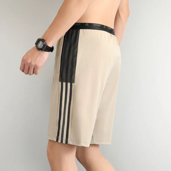 Beige athletic shorts with black side stripes and a black mesh panel on the side, worn by a person standing sideways with a wristwatch nearby.