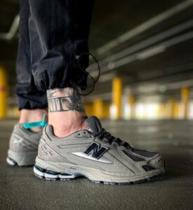Close-up of a gray New Balance sneaker on a person in a parking garage, showing a tattoo on the ankle and rolled pant leg.