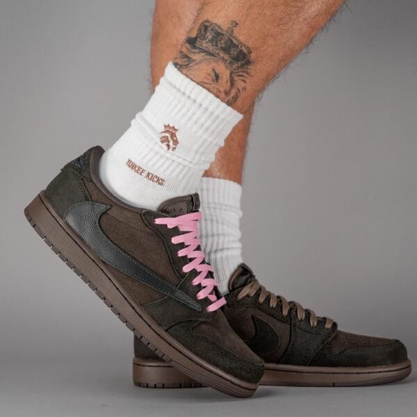 Person wearing brown suede Nike sneakers with pink laces and white crew socks, shown from the side with a tattooed leg in the background.