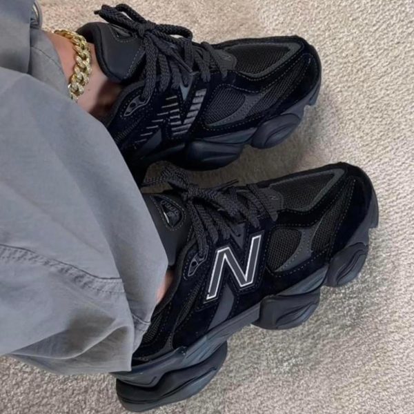 Black New Balance sneakers with thick soles, worn with gray pants and a gold chain, on a beige carpet surface.