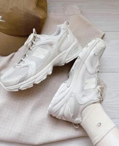 615f7c34-799f-4227-b9cc-f70f6897bd27 | UAE SPORT STORE Pair of white New Balance sneakers with mesh panels and a chunky sole, resting on beige fabric with a brown cap in the background.