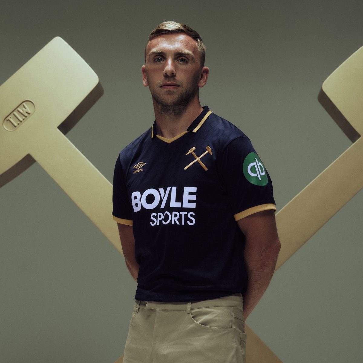 Portrait of a male athlete in a navy polo with gold trim and Boyle Sports logo, standing in front of a large gold X sculpture behind him, hands behind his back.