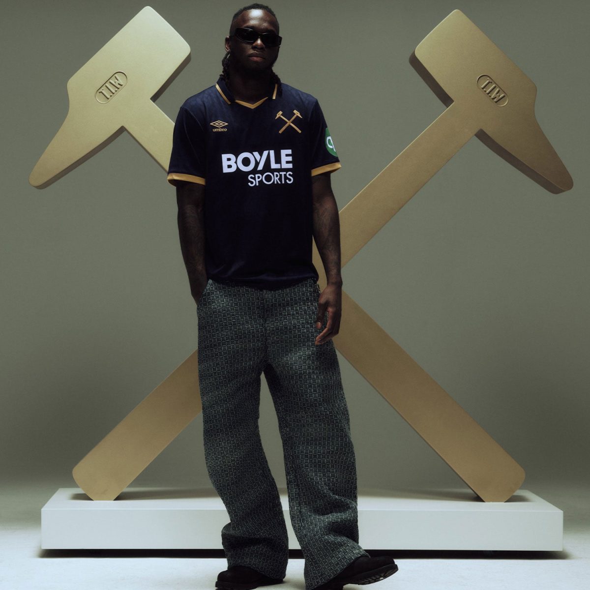Man wearing sunglasses and a dark navy sports jersey with Boyle Sports logo, posing in front of oversized gold hammers sculpture.