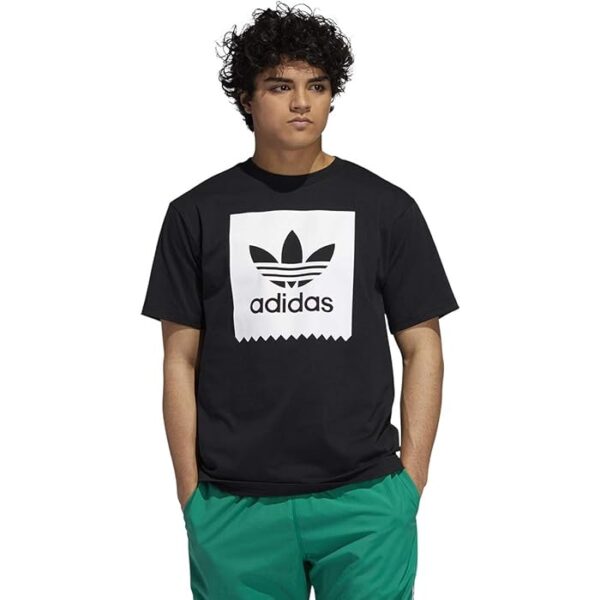 Young man in a black Adidas T-shirt with a large white logo, and teal shorts, facing slightly left with hands in pockets.