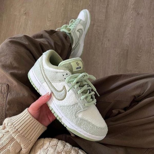 Light green Nike sneaker being held in one hand, with pastel laces and white sole, another shoe visible in the background on brown pants/wood floor. or Pastel green Nike sneaker up close, showing textured upper, mint laces, and a white-and-green sole, with a second shoe in the background.