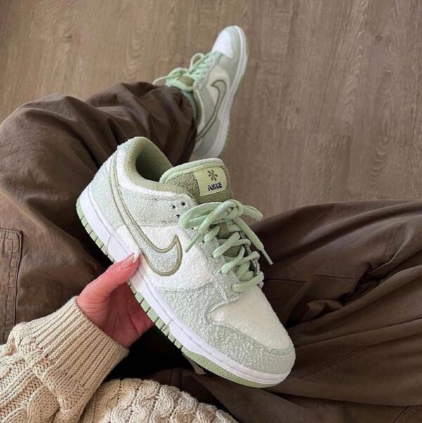Light green Nike sneaker being held in one hand, with pastel laces and white sole, another shoe visible in the background on brown pants/wood floor. or Pastel green Nike sneaker up close, showing textured upper, mint laces, and a white-and-green sole, with a second shoe in the background.