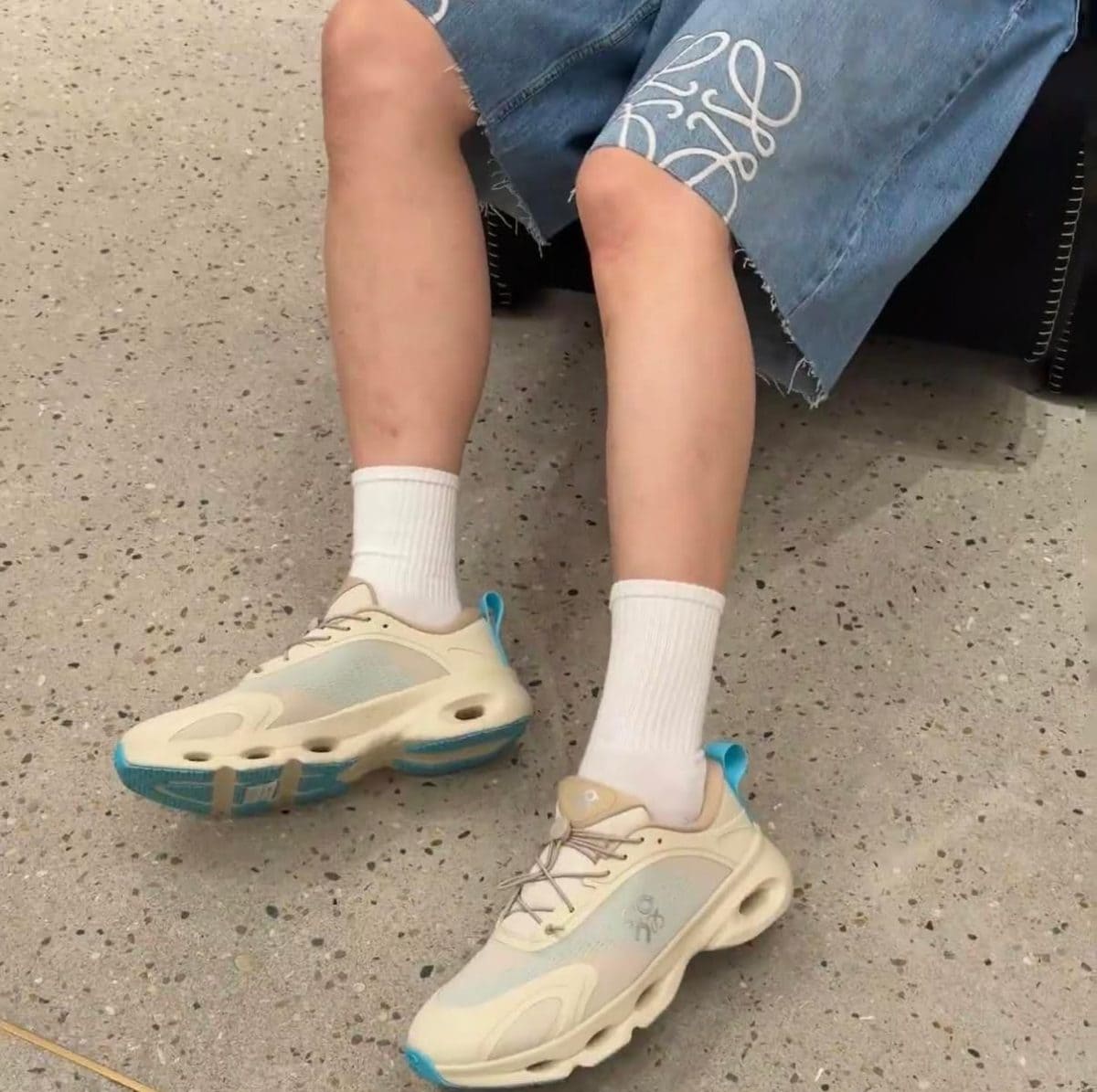 Person seated with legs shown from knee down: white socks and light-colored sneakers on a speckled floor, denim skirt visible at the knees.