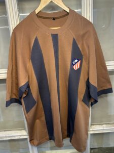 Brown oversized shirt with bold navy stripes and an Atlético Madrid crest patch on the chest.