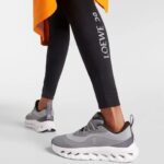 626f93fa-caac-4e09-9b0a-eda1214aa9e3 | UAE SPORT STORE Pair of grey On running sneakers with a chunky white sole, worn with black LOEWE leggings showing the LOEWE logo and an orange fabric accent in the background.