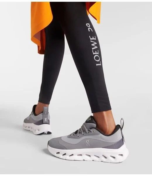 Pair of grey On running sneakers with a chunky white sole, worn with black LOEWE leggings showing the LOEWE logo and an orange fabric accent in the background.