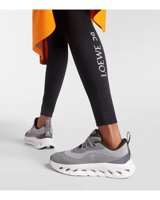 626f93fa-caac-4e09-9b0a-eda1214aa9e3 Pair of grey On running sneakers with a chunky white sole, worn with black LOEWE leggings showing the LOEWE logo and an orange fabric accent in the background.