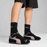 Feet in black Puma sneakers with pink side stripe, wearing black ribbed socks, against a neutral background.