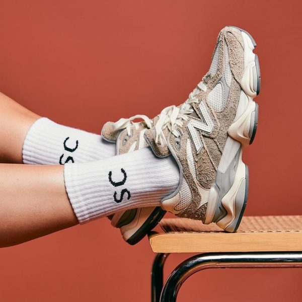 Beige chunky sneakers and white ribbed socks resting on a wooden desk against an orange background.