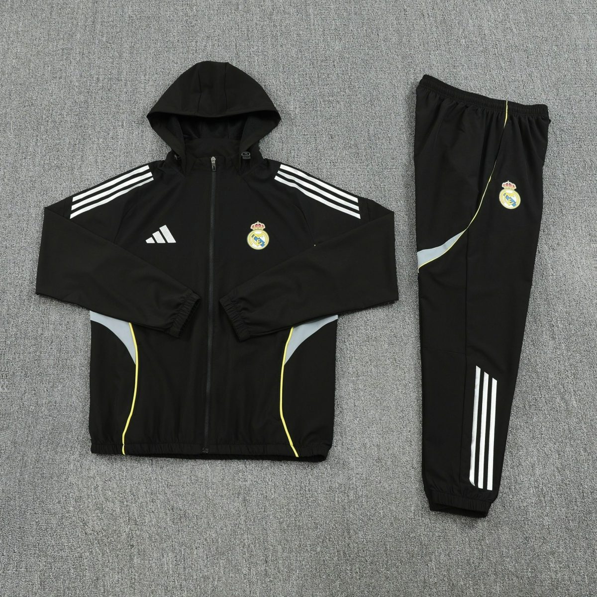 Black hooded track jacket with white three-stripe sleeves and Real Madrid crest, laid flat on a gray carpet, paired with matching black pants with white stripes.