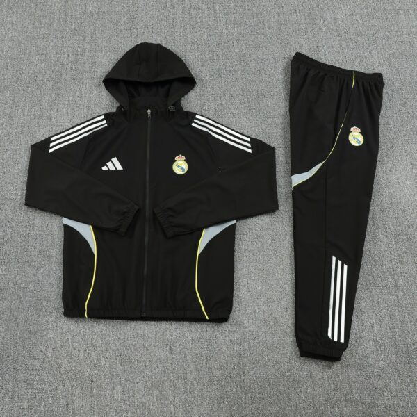 Black hooded track jacket with white three-stripe sleeves and Real Madrid crest, laid flat on a gray carpet, paired with matching black pants with white stripes.