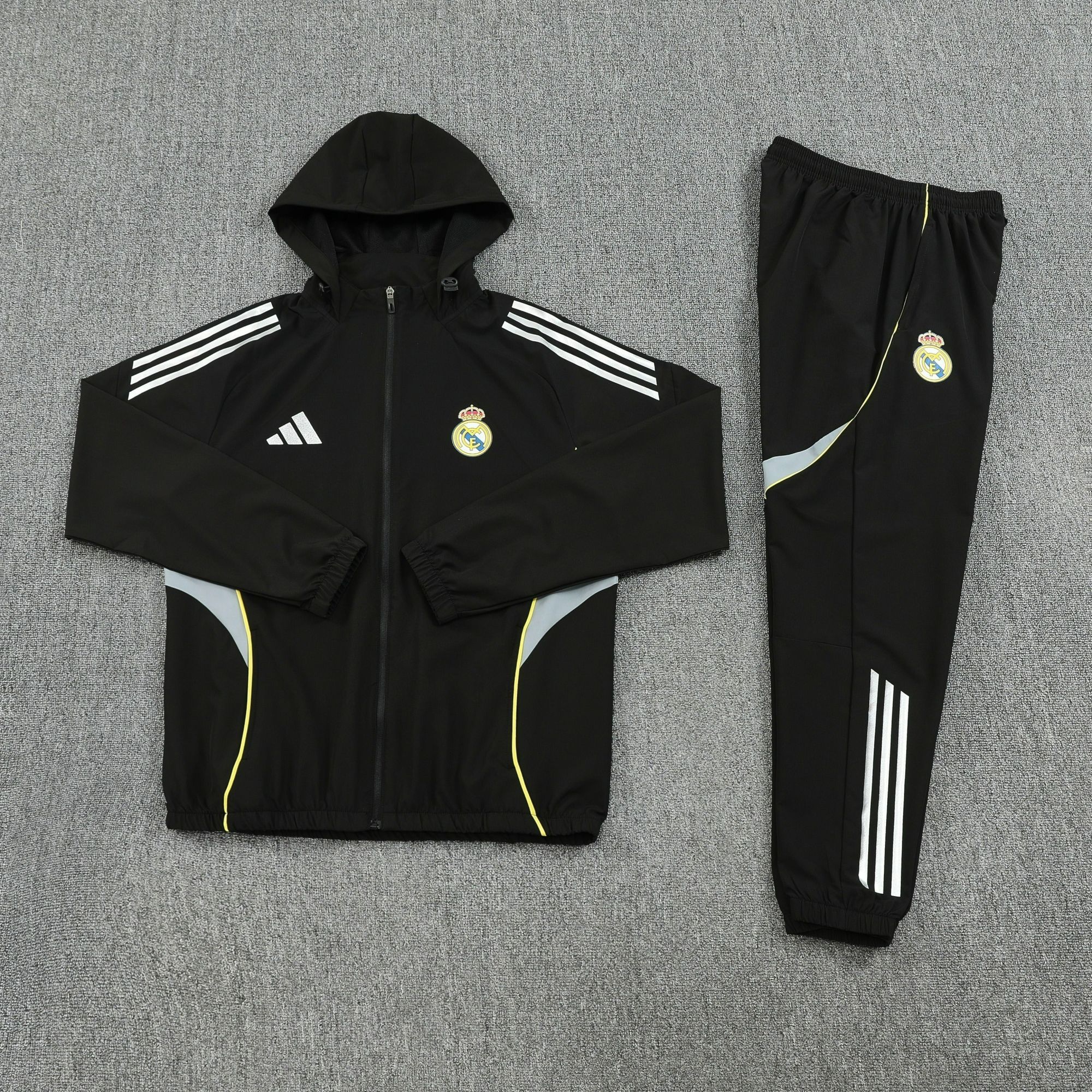63a7d877 | UAE SPORT STORE Black hooded track jacket with white three-stripe sleeves and Real Madrid crest, laid flat on a gray carpet, paired with matching black pants with white stripes.