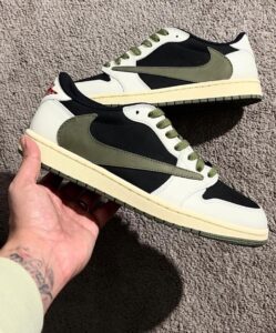 Pair of white, black, and olive-green sneakers with cream soles resting on a carpet; one shoe held in a hand.
