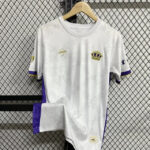 White soccer jersey with purple side panels and cuffs, gold crown crest on the chest, hanging on a wooden hanger against a metal grid background.