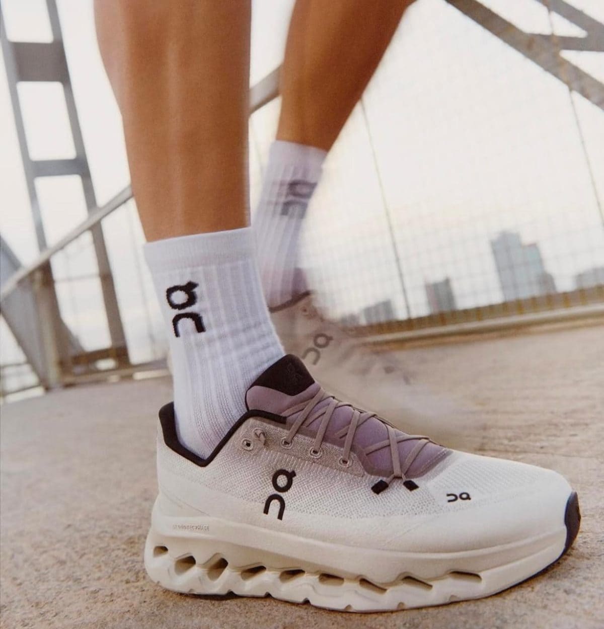 Close-up of a white On running sneaker and white socks with the On logo, standing on a bridge with a city skyline in the background.