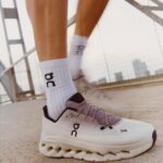 645f3e8e-718e-4095-ae1b-acde4a3243fd | UAE SPORT STORE Close-up of a white On running sneaker and white socks with the On logo, standing on a bridge with a city skyline in the background.