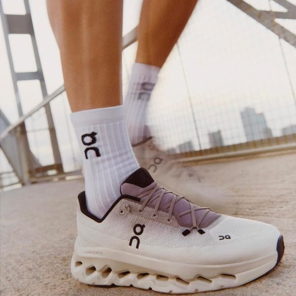 Close-up of a white On running sneaker and white socks with the On logo, standing on a bridge with a city skyline in the background.