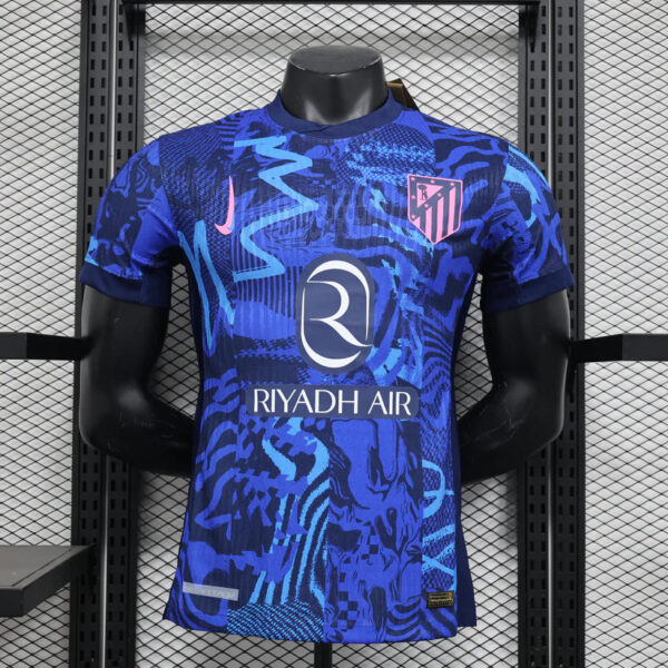 Blue Atlético Madrid jersey with bold teal and blue abstract pattern, Riyadh Air sponsor front center, crest on chest, displayed on a mannequin on a rack.