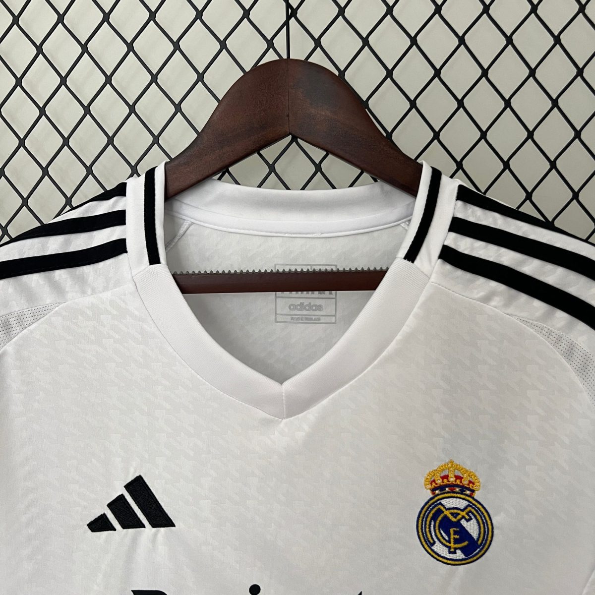 White Real Madrid soccer jersey on a wooden hanger, with the adidas logo and Real Madrid crest visible, set against a diamond-pattern chain-link fence.